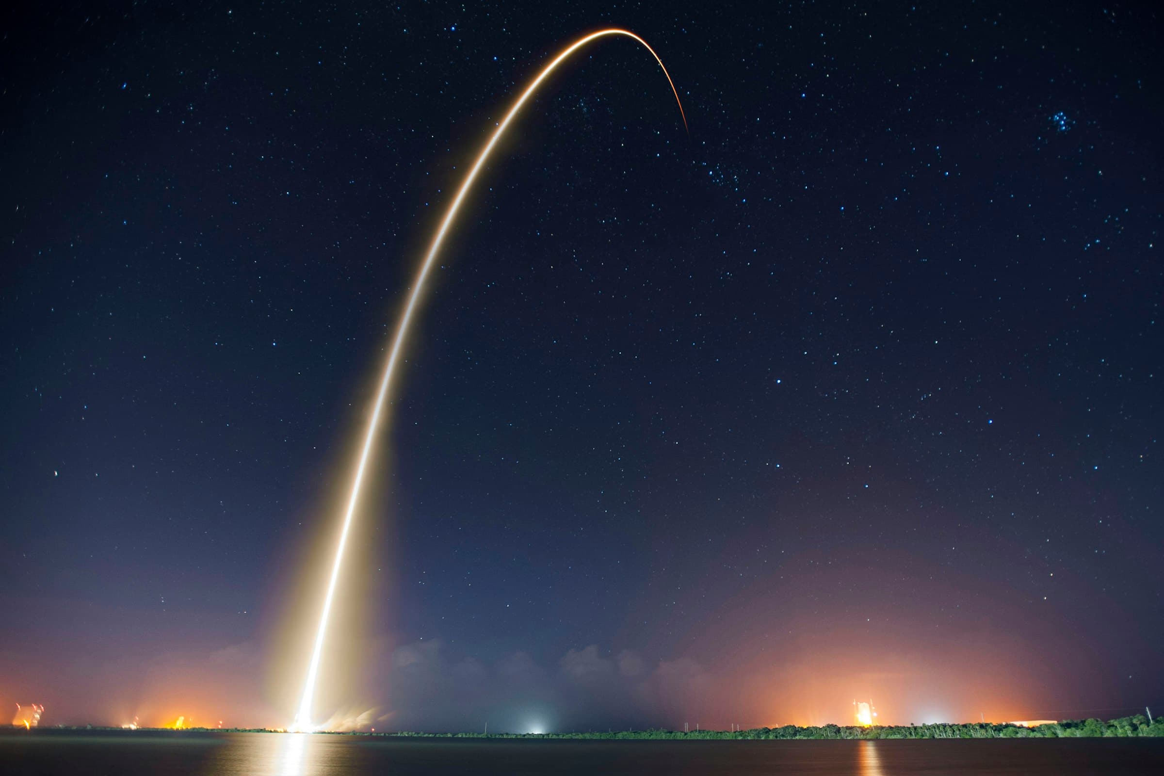 A rocket launch, seen as a long golden light trail arcing across a starry night sky